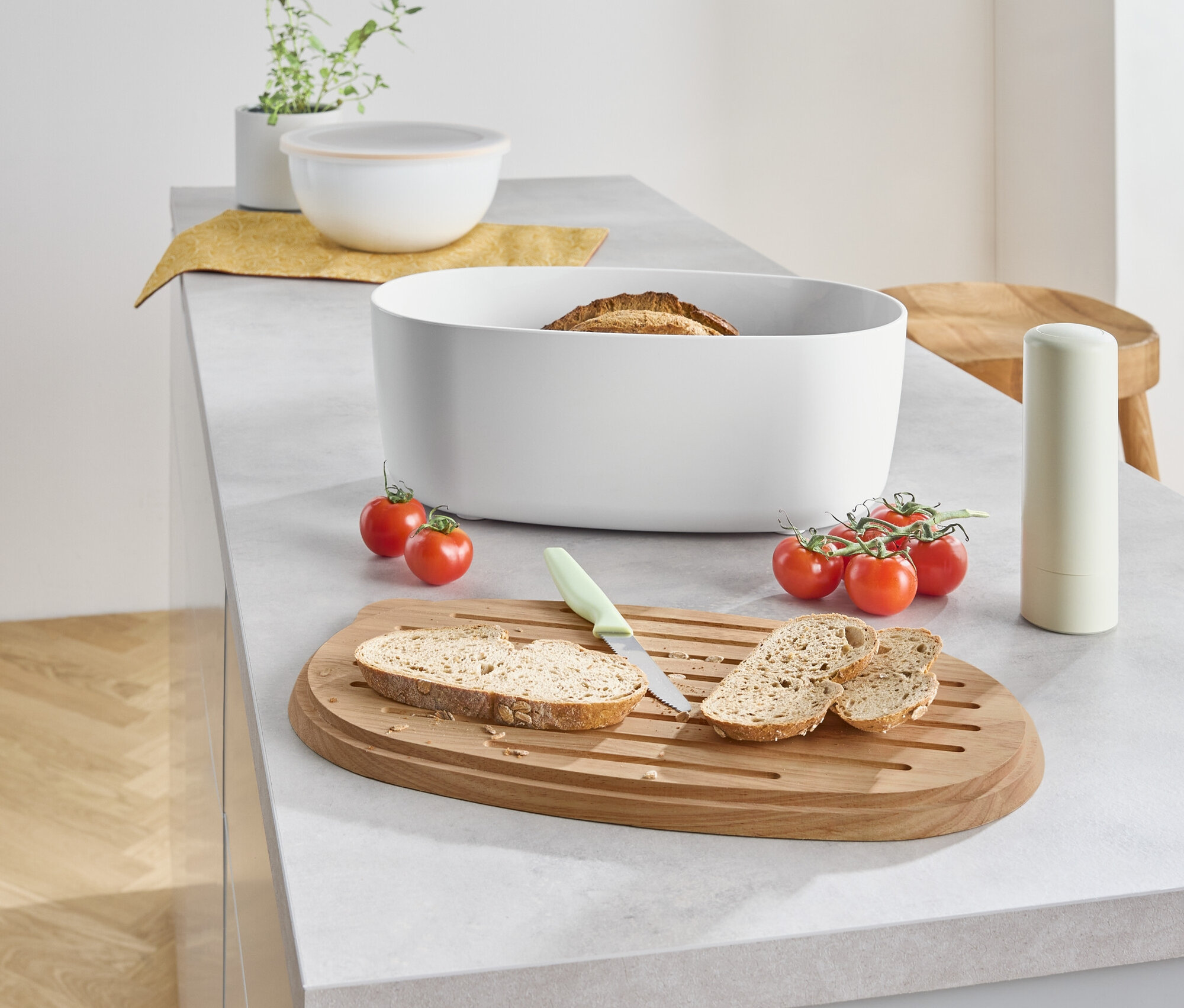 Na kuchyňské lince leží chléb v bílé nádobě, vedle něj je nakrájený chléb na dřevěném prkénku s nožem a cherry rajčaty.