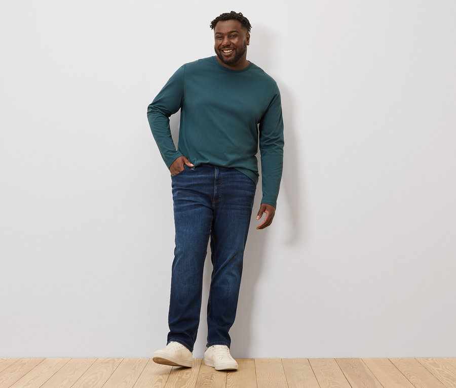 Muž stojí a má na sobě zelené tričko s dlouhým rukávem a džíny. Na sobě má 2 ks trika s dlouhým rukávem.