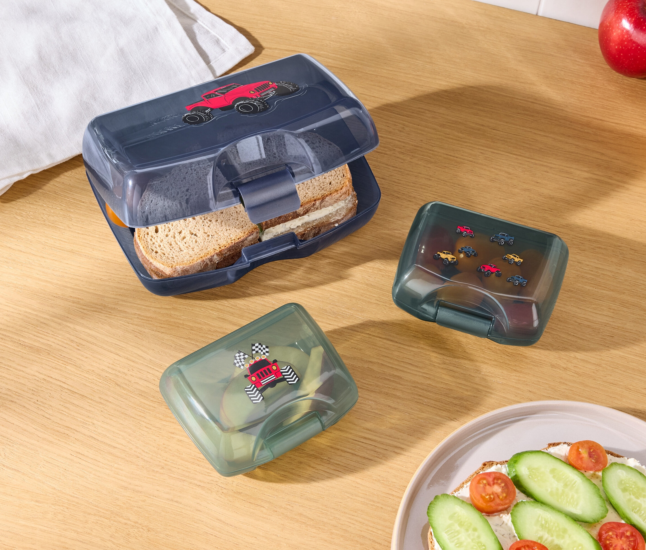 Na dřevěném stole leží otevřený modrý box na svačinu s obrázkem červeného auta, dva zelené boxy na svačinu s obrázky aut, jablko, pomeranč, bílá utěrka a talíř s chlebíčkem.