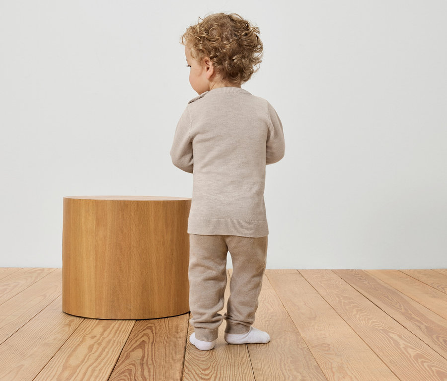 Batole stojí zády ke kameře v béžovém svetru a kalhotách vedle dřevěného stolku.
