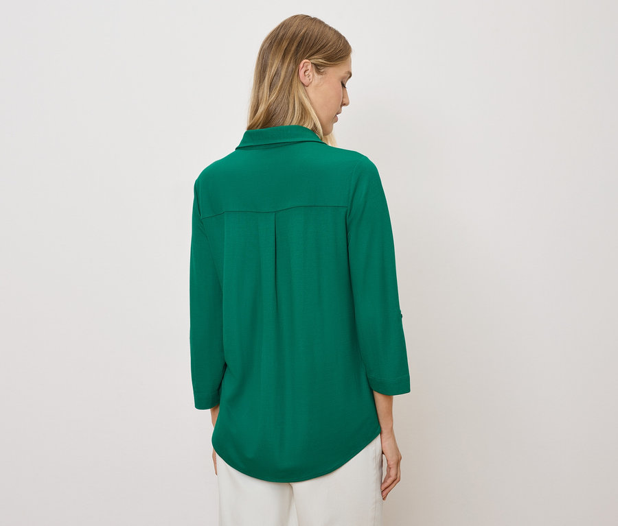 Žena zády k fotoaparátu má na sobě zelenou košili a bílé kalhoty.