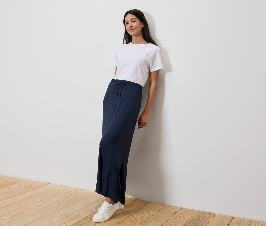 Žena se opírá o zeď, má na sobě bílé tričko, dlouhou tmavě modrou sukni a bílé tenisky.