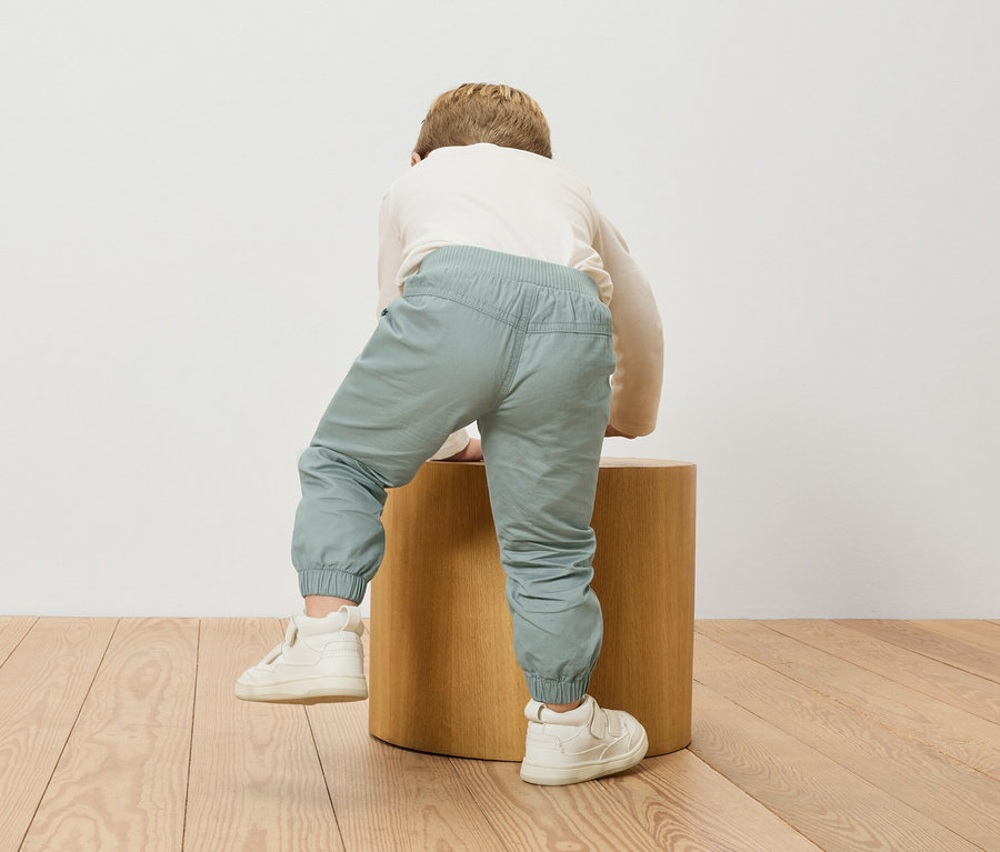Dítě leze na dřevěnou stoličku, má na sobě blůzu s dlouhým rukávem a světle modré kalhoty.