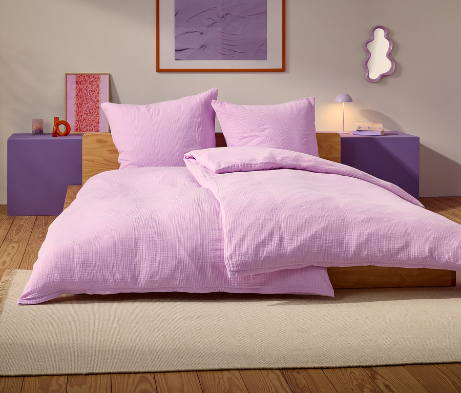 Fialová postel se dvěma polštáři a přikrývkou stojí na béžovém koberci v ložnici.