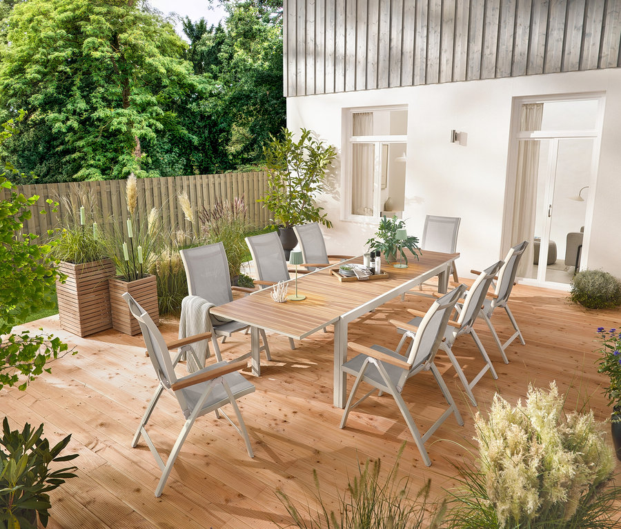 Rozkládací hliníkový zahradní stůl »Liska« o délce cca 200–300 cm stojí na dřevěné terase a je obklopen šesti vysokými křesly »Liska« s nastavitelným opěradlem. Jsou zde také 2 truhlíky »Tinus« a 5 zahradních LED pochodní.