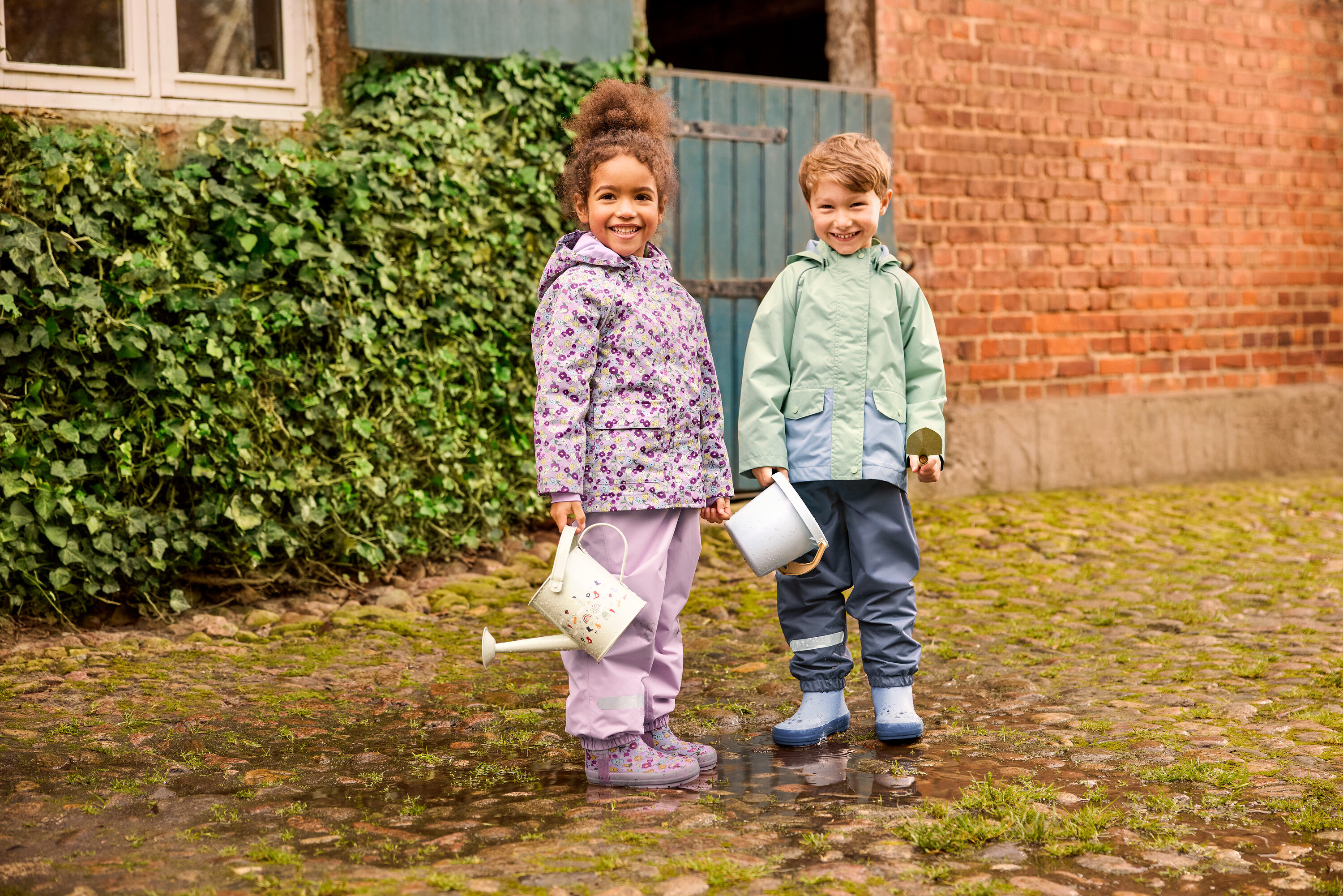 Dvě děti stojí venku: dívka v květinové dětské pláštěnce a květinových dětských holínkách a chlapec v zelené dětské pláštěnce a modrých dětských holínkách. Oba mají na sobě dětské kalhoty do deště.