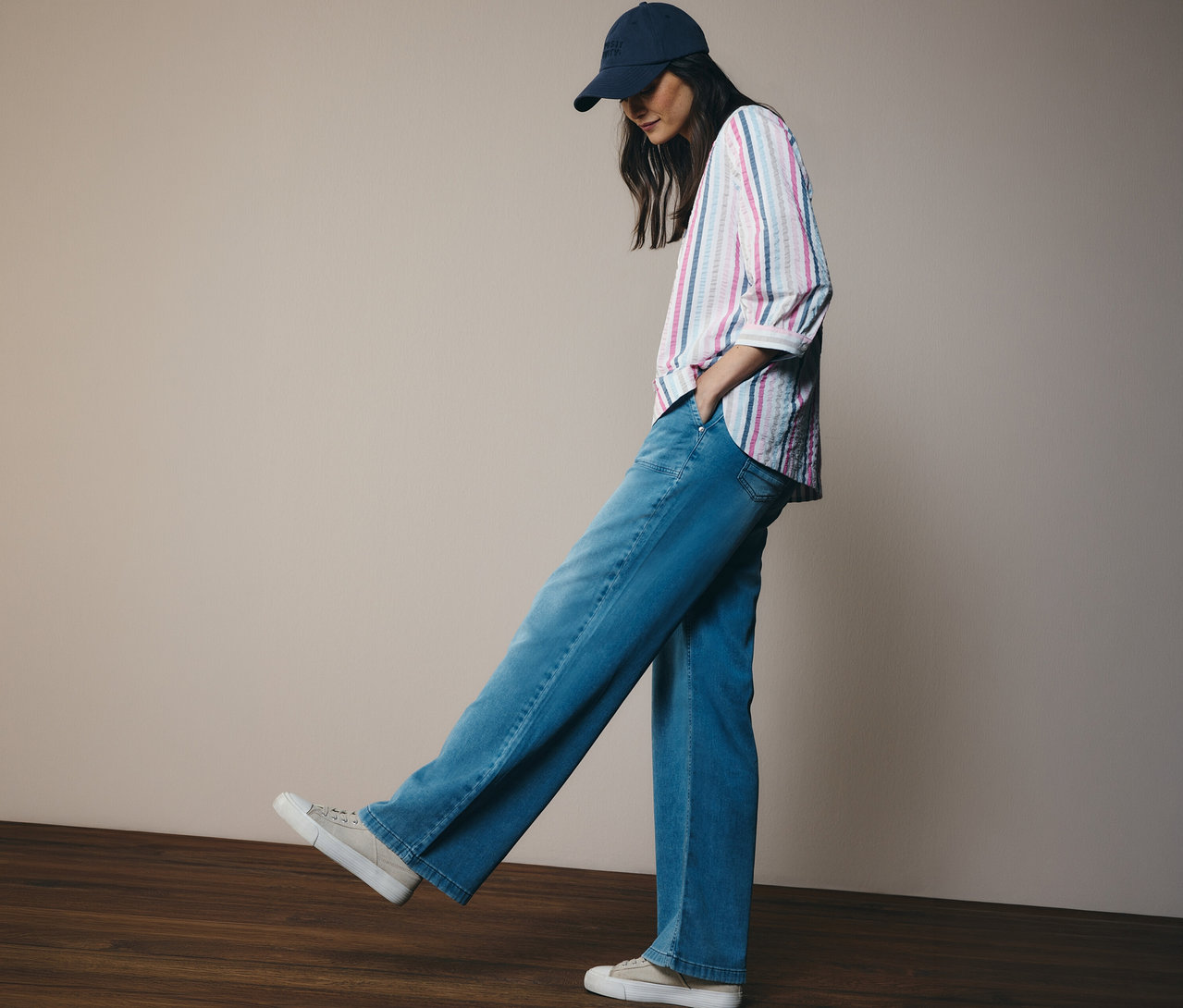 Žena v džínách a baseballové čepici má na sobě košilovou halenku CECIL s proužky. Stojí na dřevěné podlaze.