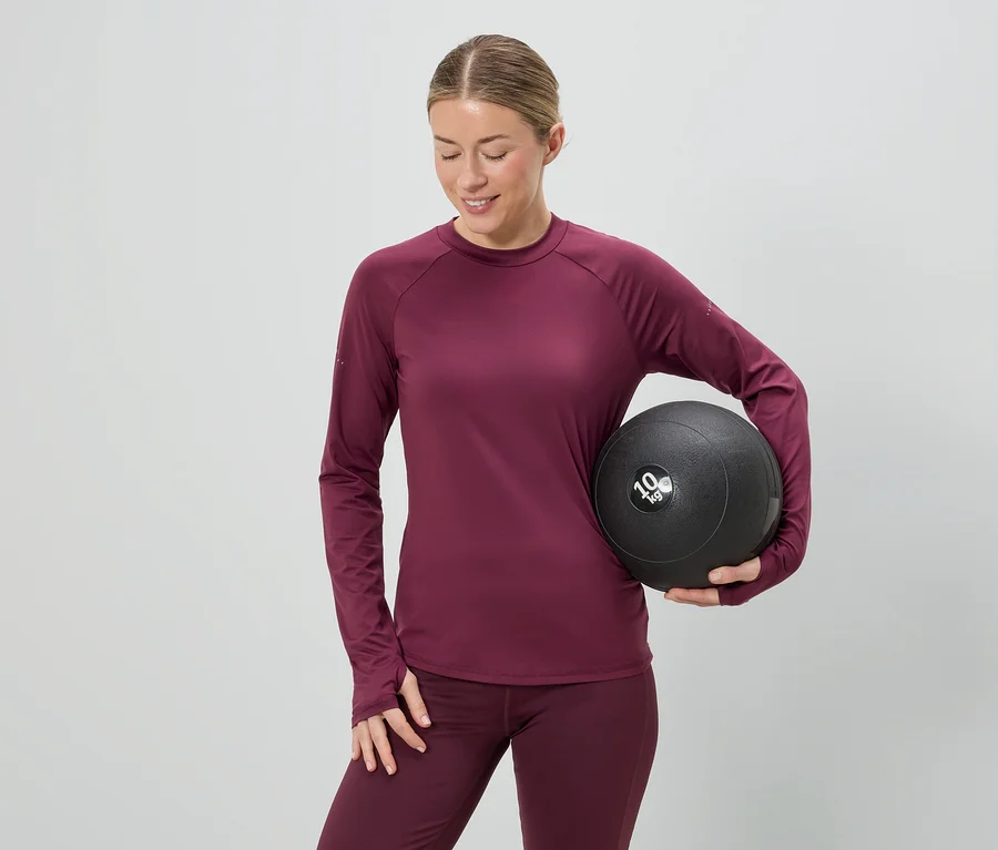 Žena v bordovém sportovním oblečení drží 10 kg medicinbal na šedém pozadí.