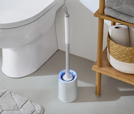 V koupelně stojí bílá toaleta vedle WC štětky s modrým světlem a dřevěného regálu s ručníky a toaletním papírem.