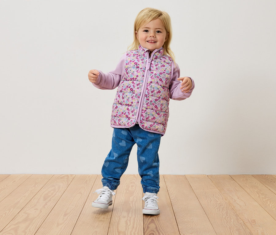 Malá blondýnka stojí na dřevěné podlaze a má na sobě květinovou vestu, fialový top a džíny.