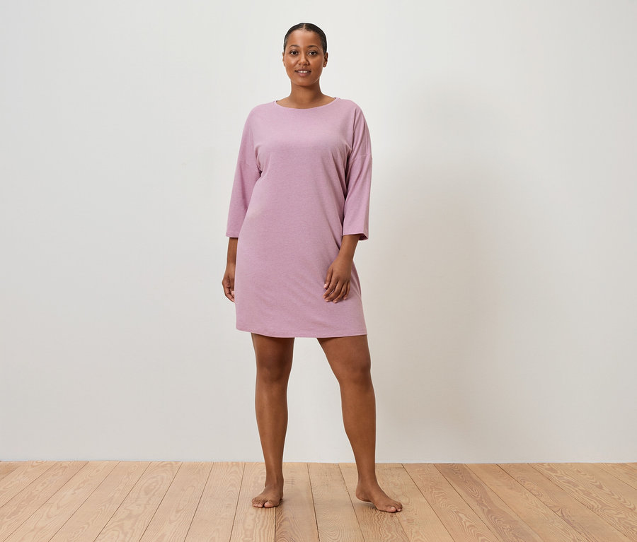 Žena stojí bosá před zdí a má na sobě růžové šaty s krátkým rukávem.