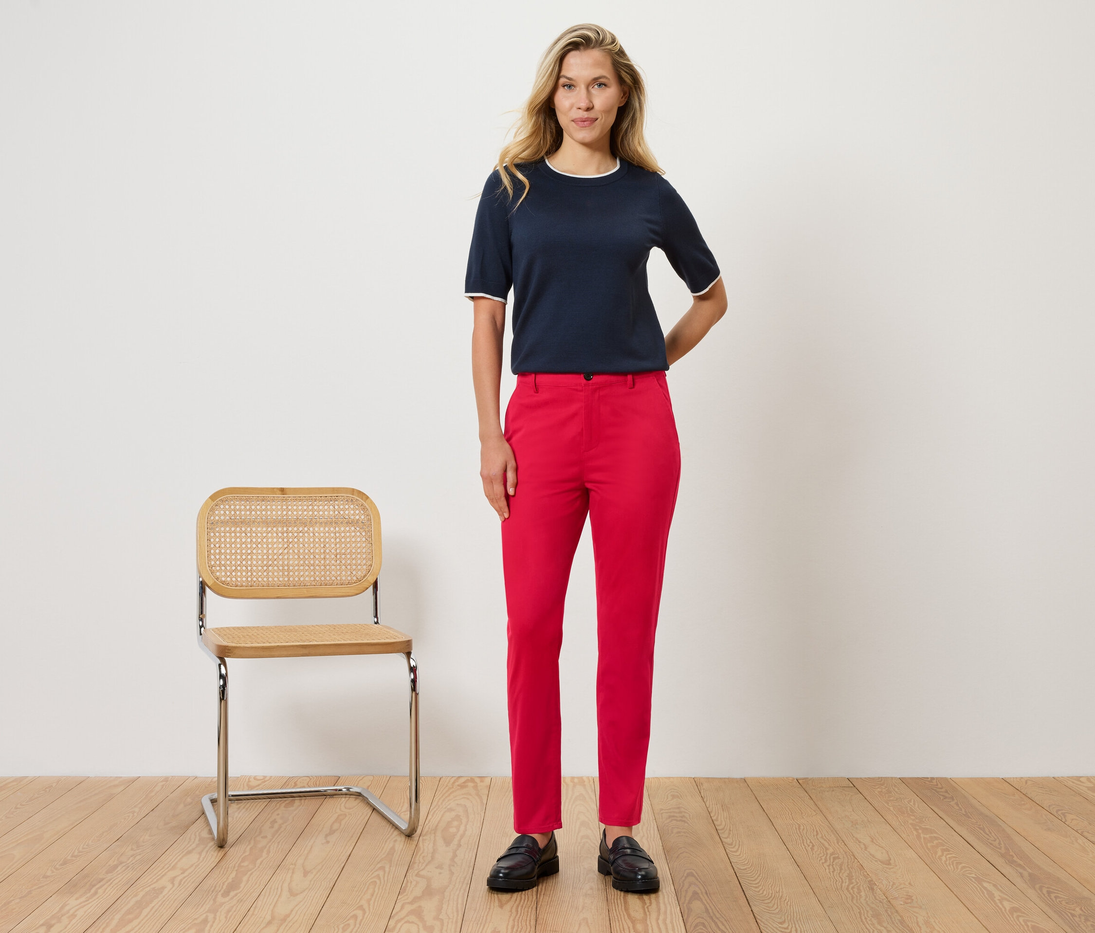 Žena stojí před zdí vedle židle. Má na sobě tmavě modrou blůzu s bílými detaily a červené kalhoty.