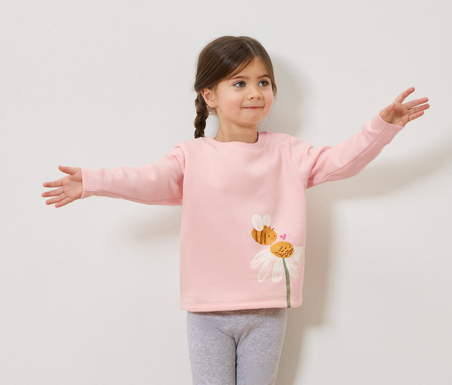 Dívka se zapletenými vlasy v růžovém svetru s potiskem včely a sedmikrásky, šedé legíny, s nataženýma rukama.