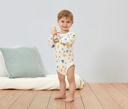 Usmívající se chlapec v body s listovým vzorem drží chrastítko. V pozadí je vidět polštář a futon.