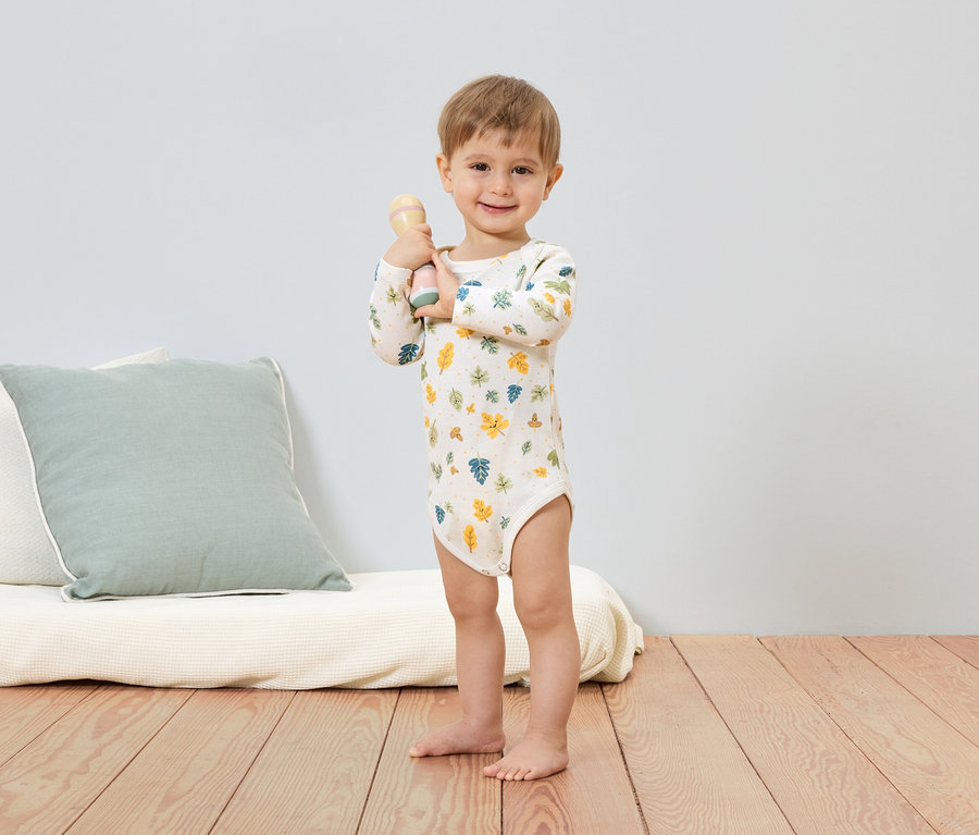 Usmívající se chlapec v body s listovým vzorem drží chrastítko. V pozadí je vidět polštář a futon.