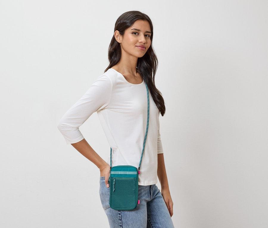 Žena s tmavě hnědými vlasy nosí zelenou tašku přes rameno s tyrkysovým popruhem, bílé tričko s dlouhým rukávem a džíny.