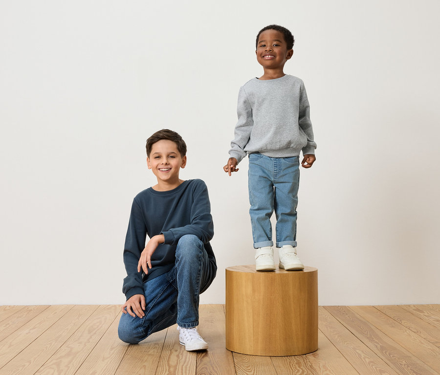 Dva chlapci pózují: jeden klečí v tmavě modré mikině a džínách, druhý stojí na dřevěné stoličce v šedé mikině a džínách.