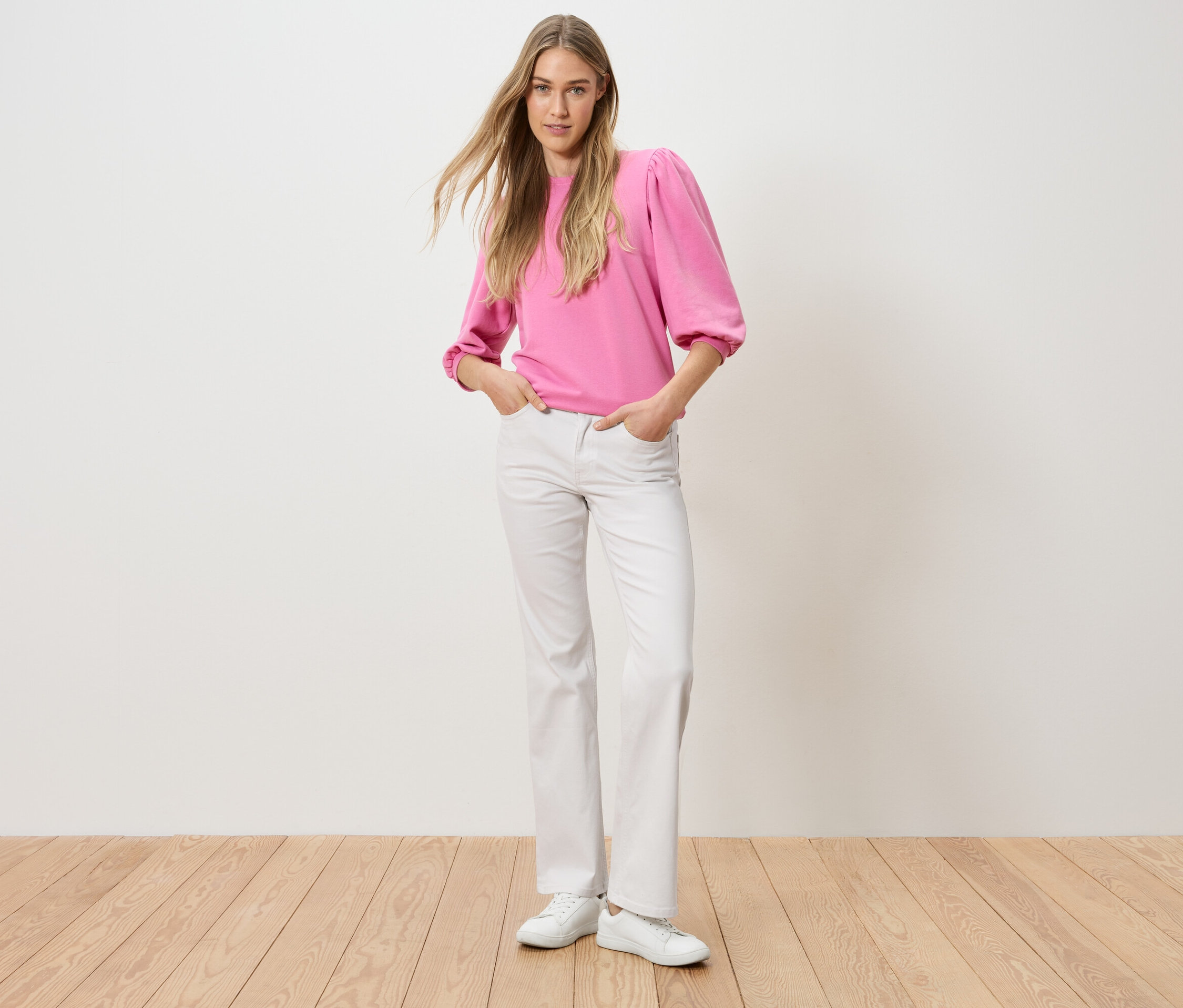 Žena stojí s rukama v kapsách kalhot. Má na sobě růžový top a bílé džíny.