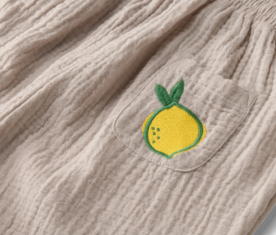 Detail béžových látkových kalhot s elastickým pasem a našitou kapsou s výšivkou žlutého citronu.