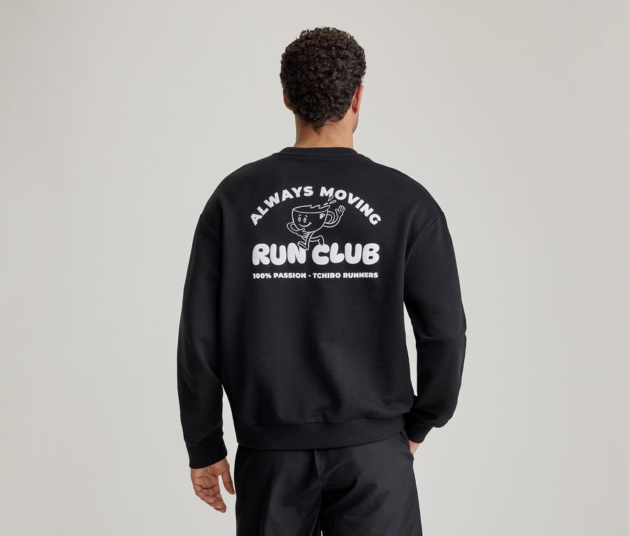 Muž zezadu v černé mikině s bílým potiskem 'ALWAYS MOVING RUN CLUB' a kresleným běžícím šálkem kávy na zádech.