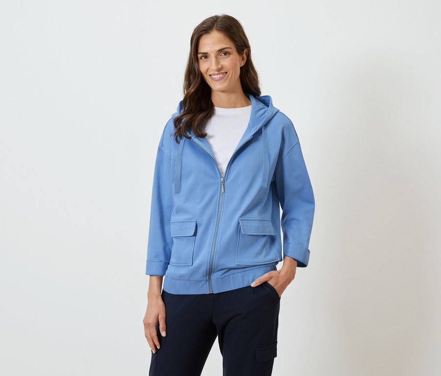 Žena s dlouhými hnědými vlasy se usmívá v modré mikině s kapucí a tmavých kalhotách.