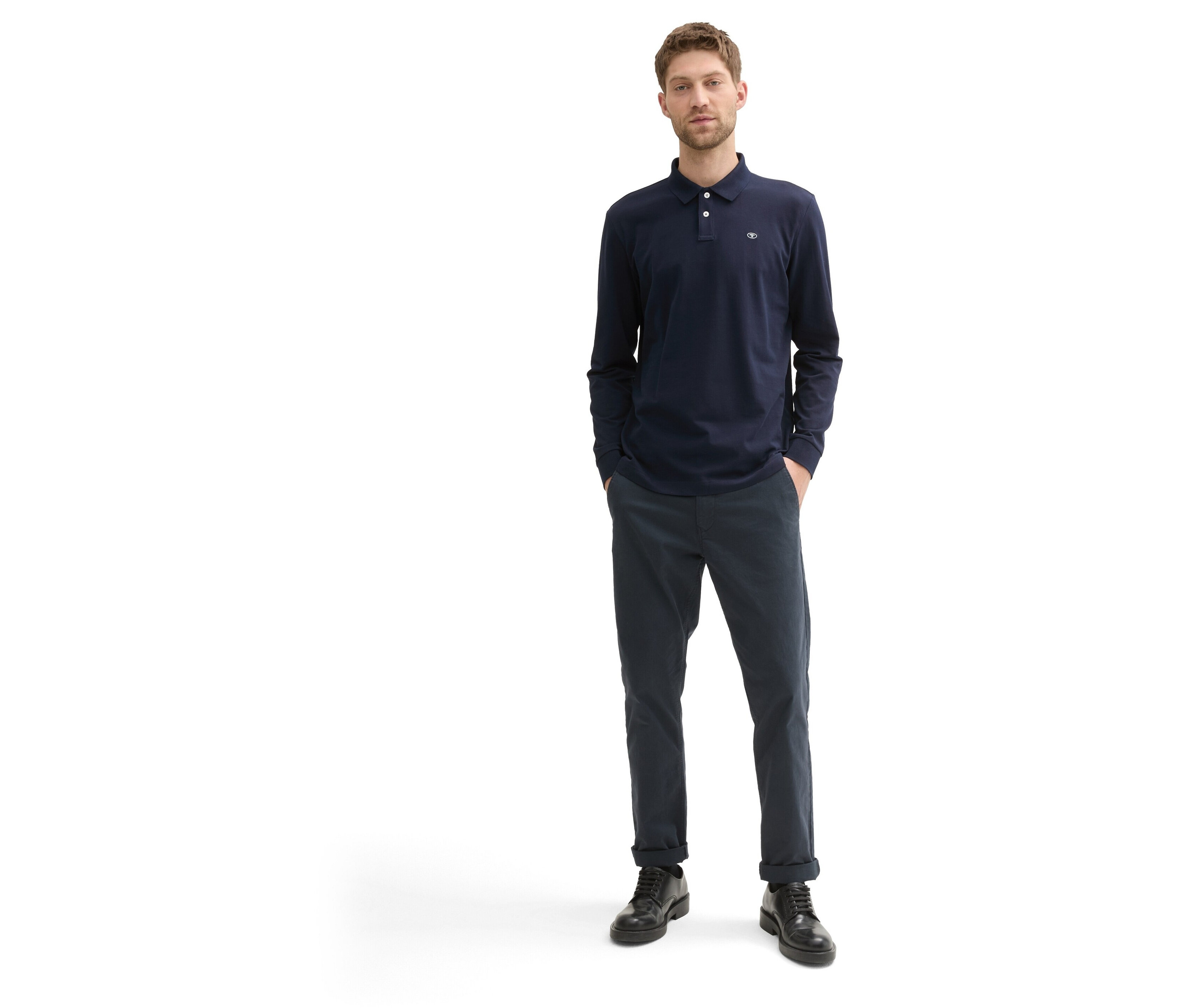 Muž stojí a má na sobě tmavě modré polo triko s dlouhým rukávem, tmavě modré kalhoty a černé boty.