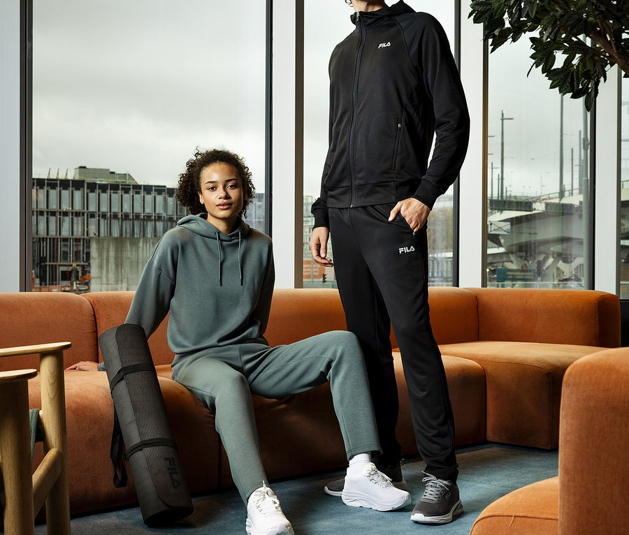 Mladá žena sedí srolovanou podložkou na jógu vedle sebe na oranžové pohovce, vedle ní stojí mladý muž. Oba mají na sobě teplákové soupravy a tenisky.