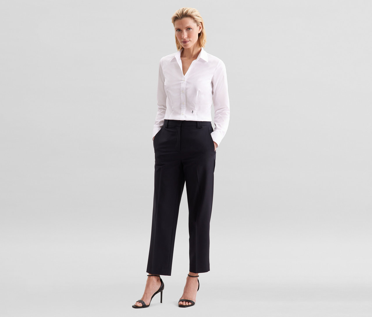 Žena s blond vlasy stojí s rukama v kapsách černých kalhot. Má na sobě bílou halenku slim fit non iron Seidensticker a černé sandály na vysokém podpatku.