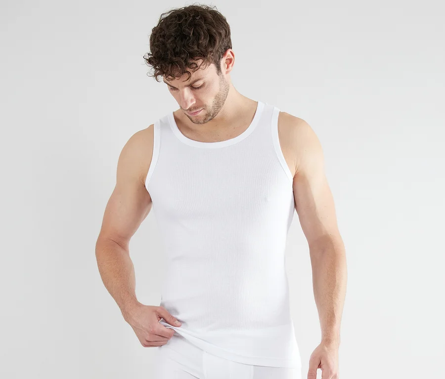 Muž v bílém, žebrovaném nátělníku Tom Tailor Montana. Dívá se dolů a drží lem nátělníku.