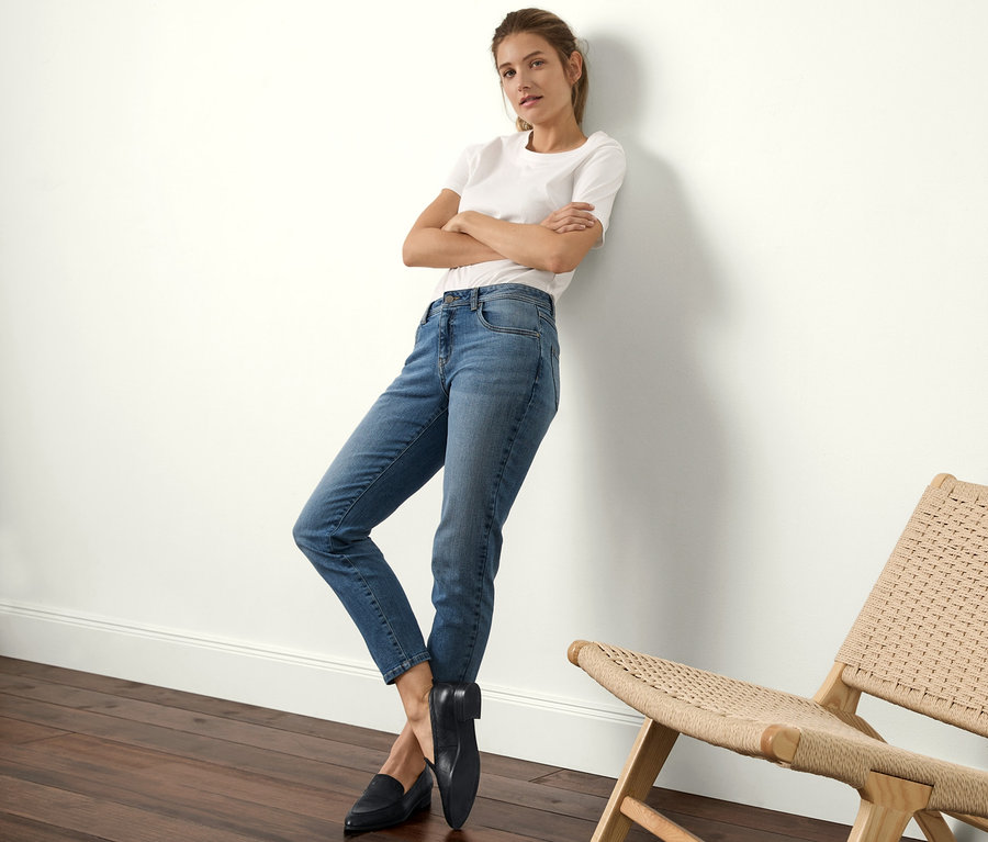 Žena opřená o zeď má na sobě džíny slimfit, bílé tričko a černé mokasíny.