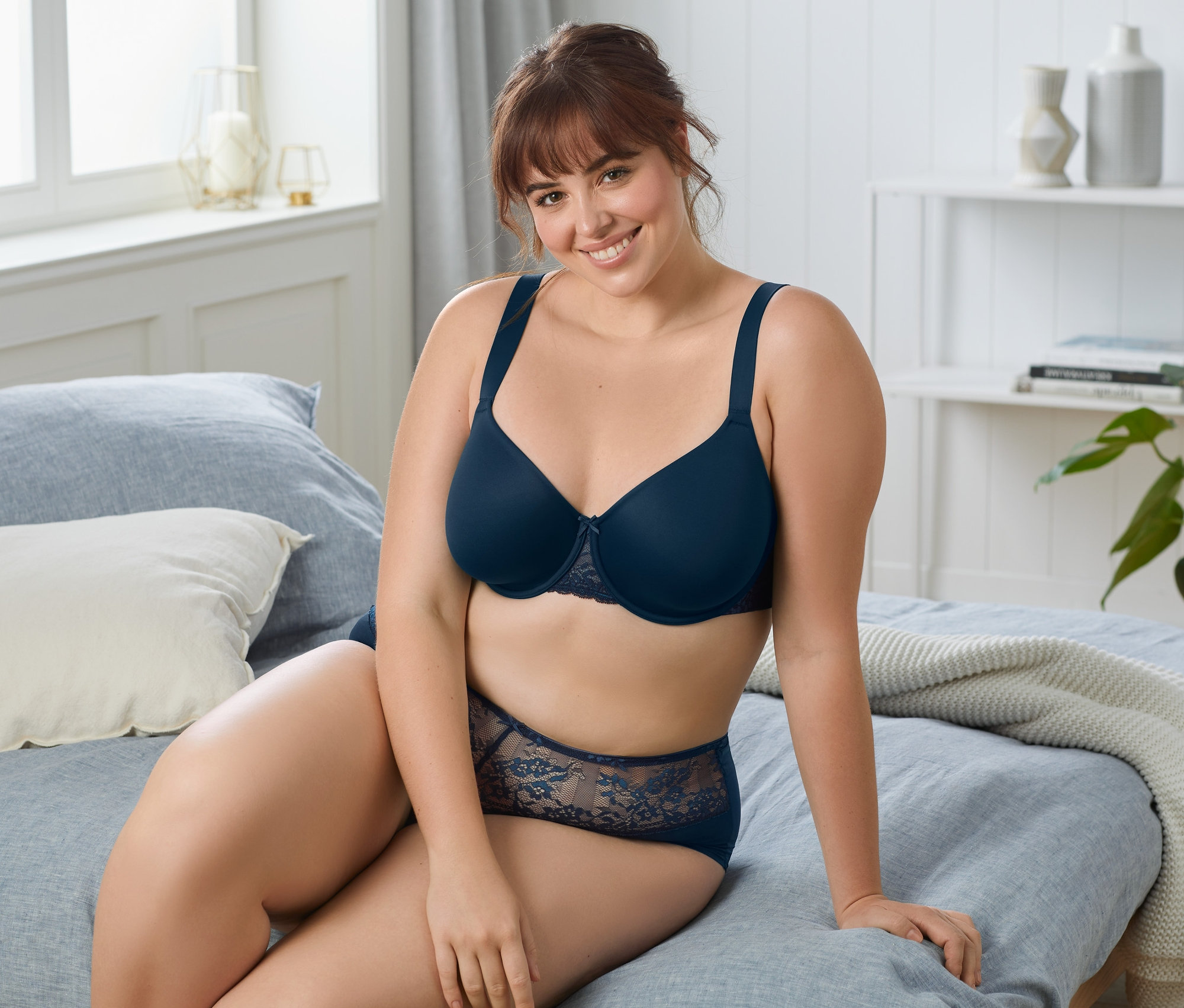 Žena sedí na posteli a má na sobě tmavě modrou podprsenku s kosticemi.