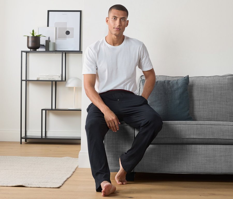 Muž sedí na šedé pohovce a má na sobě bílé tričko a pohodlné kalhoty.