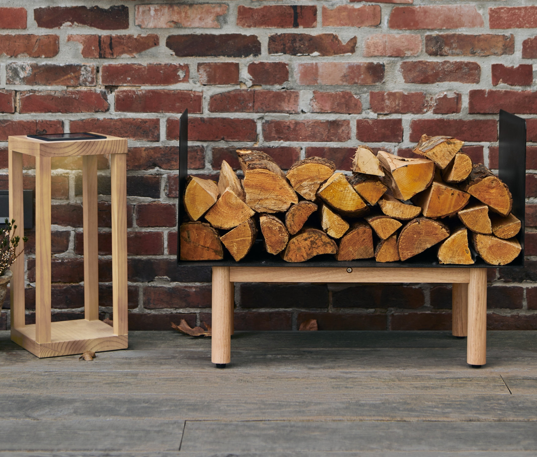 Na cihlové zdi visí věnec, pod ním stojí regál na krbové dřevo plný polen a vedle něj dřevěná solární lucerna.