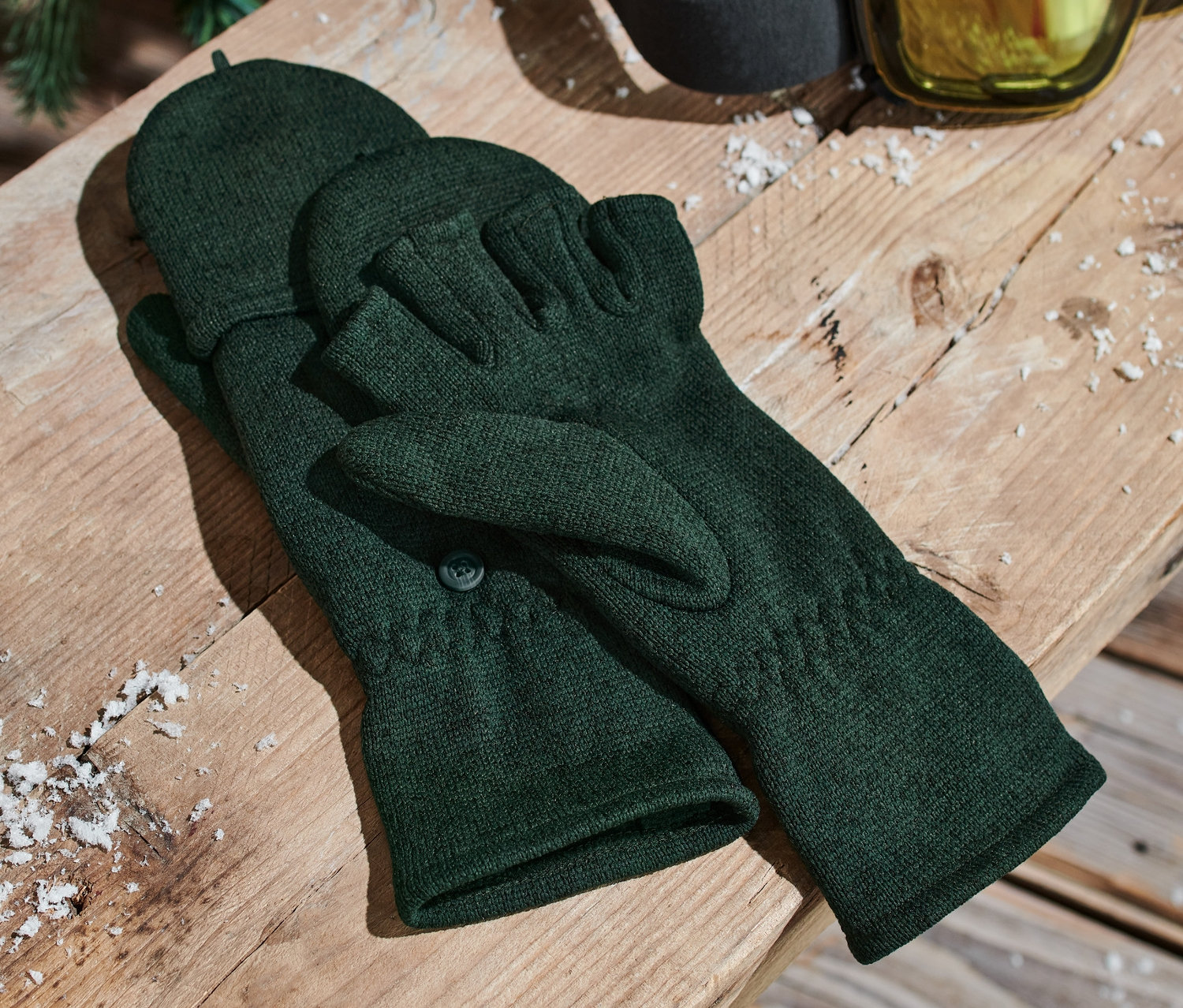 Zelené pletené flísové odklápěcí palčáky leží na dřevěné lavici vedle lyžařských brýlí.