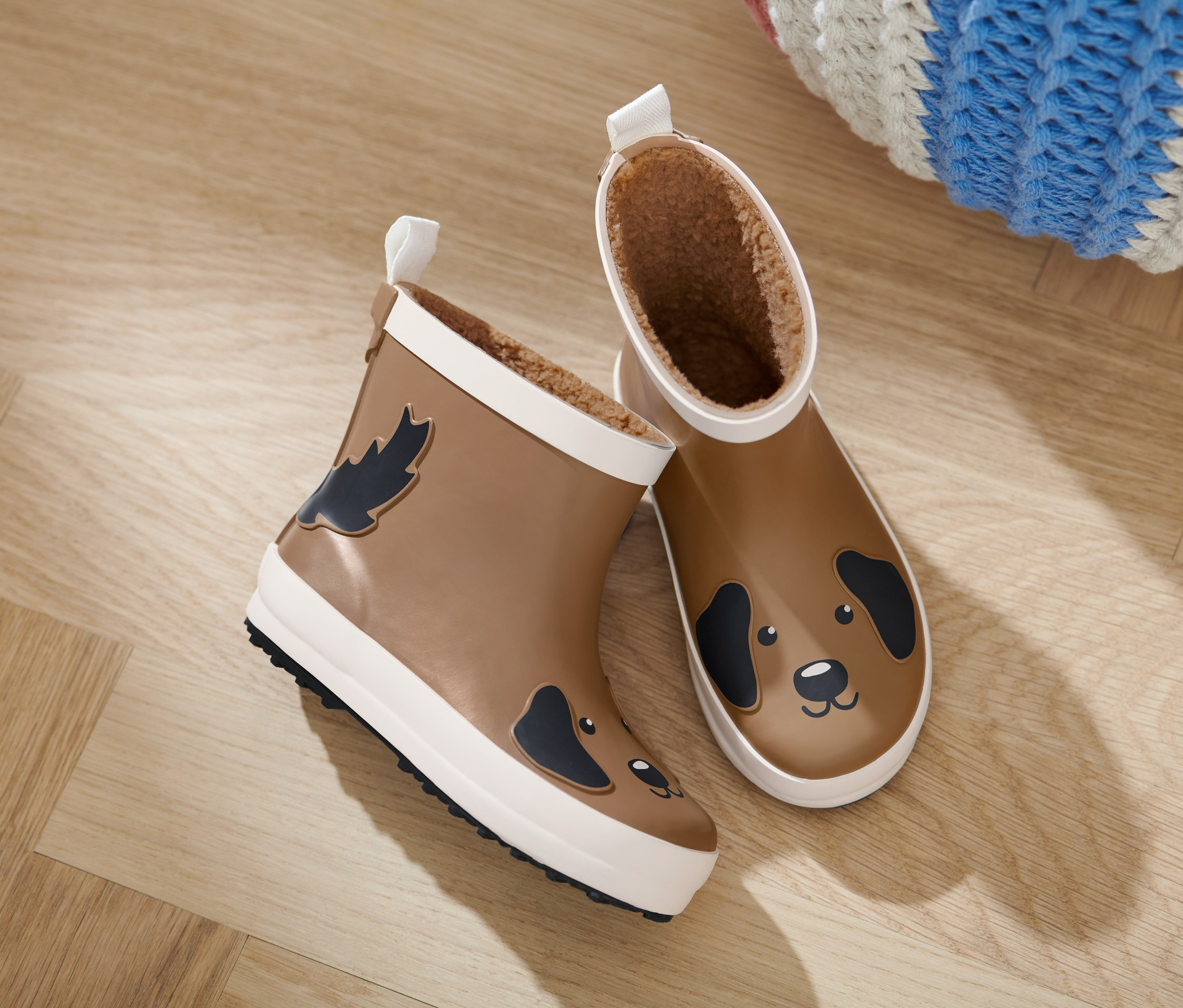 Dvoje dětské holínky Chelsea na dřevěné podlaze. Jedna bota má na straně obrázek listu a druhá obrázek psa.