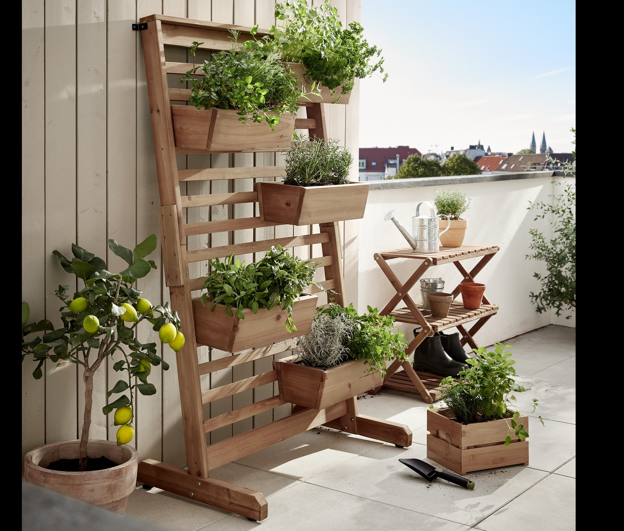 Na terase stojí skládací police »Tinus« a vertikální záhon »Tinus« s truhlíky, oba osázené zelenými rostlinami.