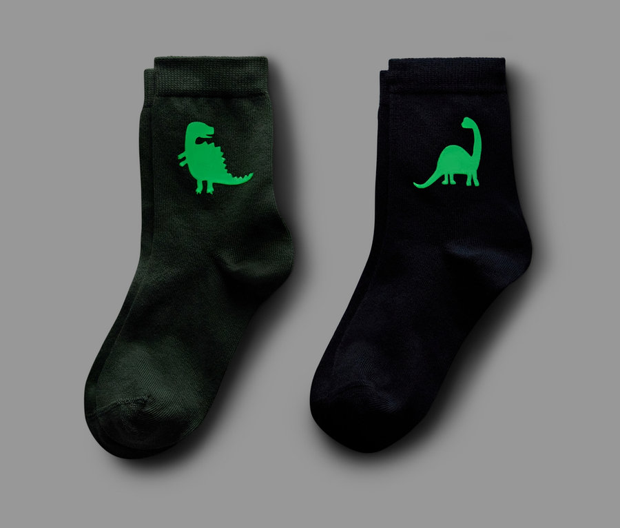Dva páry ponožek: jeden tmavě zelený s motivem dinosaura, druhý černý s motivem dinosaura. Oba páry ponožek svítí ve tmě.