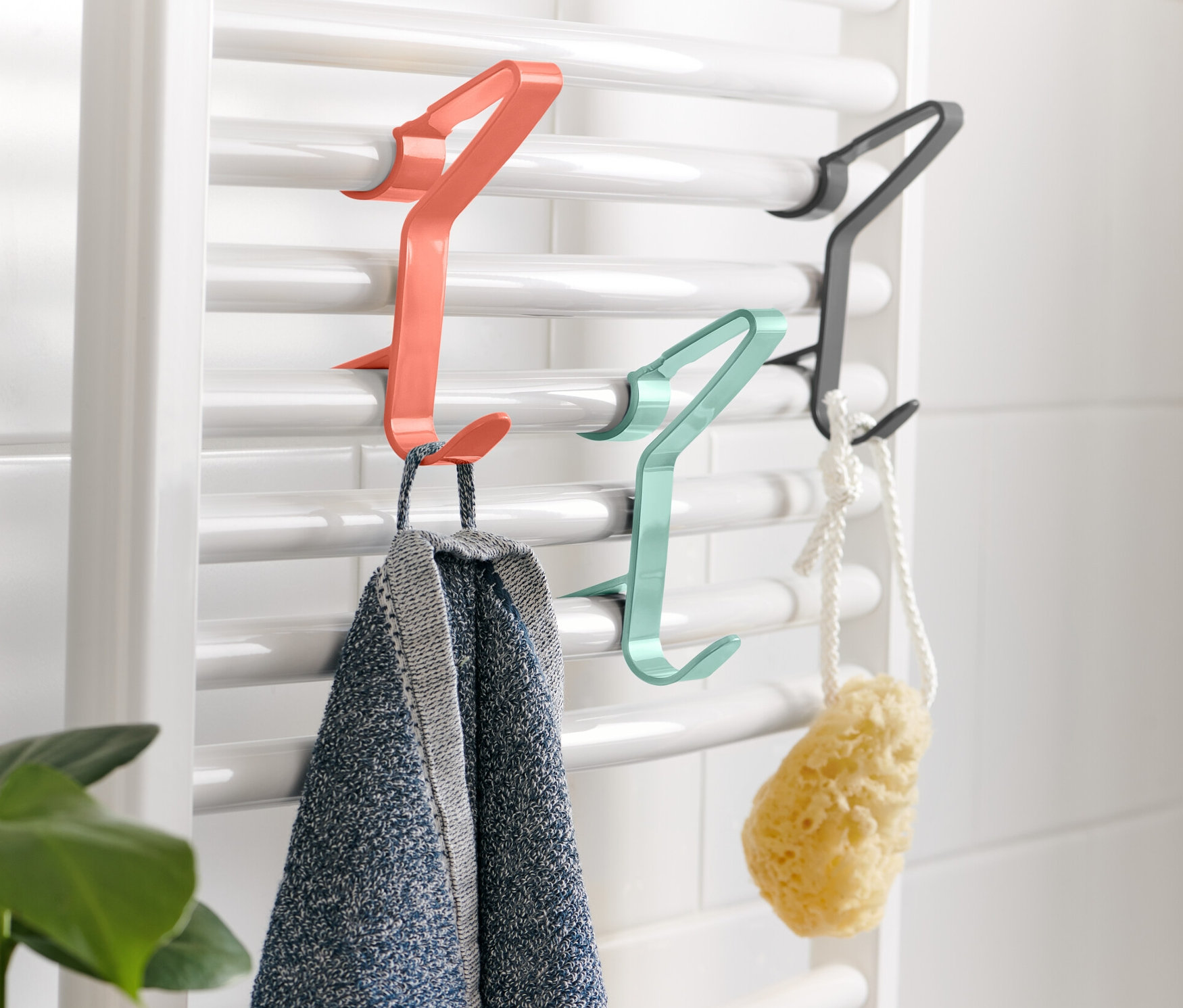 Na bílém radiátoru jsou zavěšeny 3 háčky na topná tělesa, na kterých visí ručník a houba.