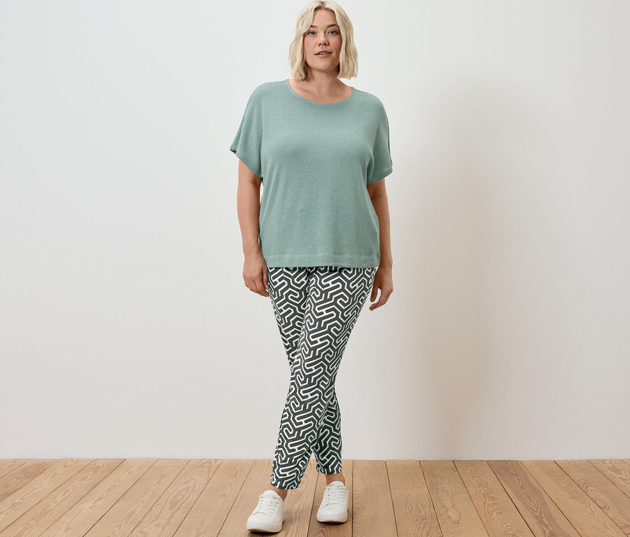 Žena stojí a má na sobě zelené strukturované tričko a zelené žerzejové kalhoty s potiskem.