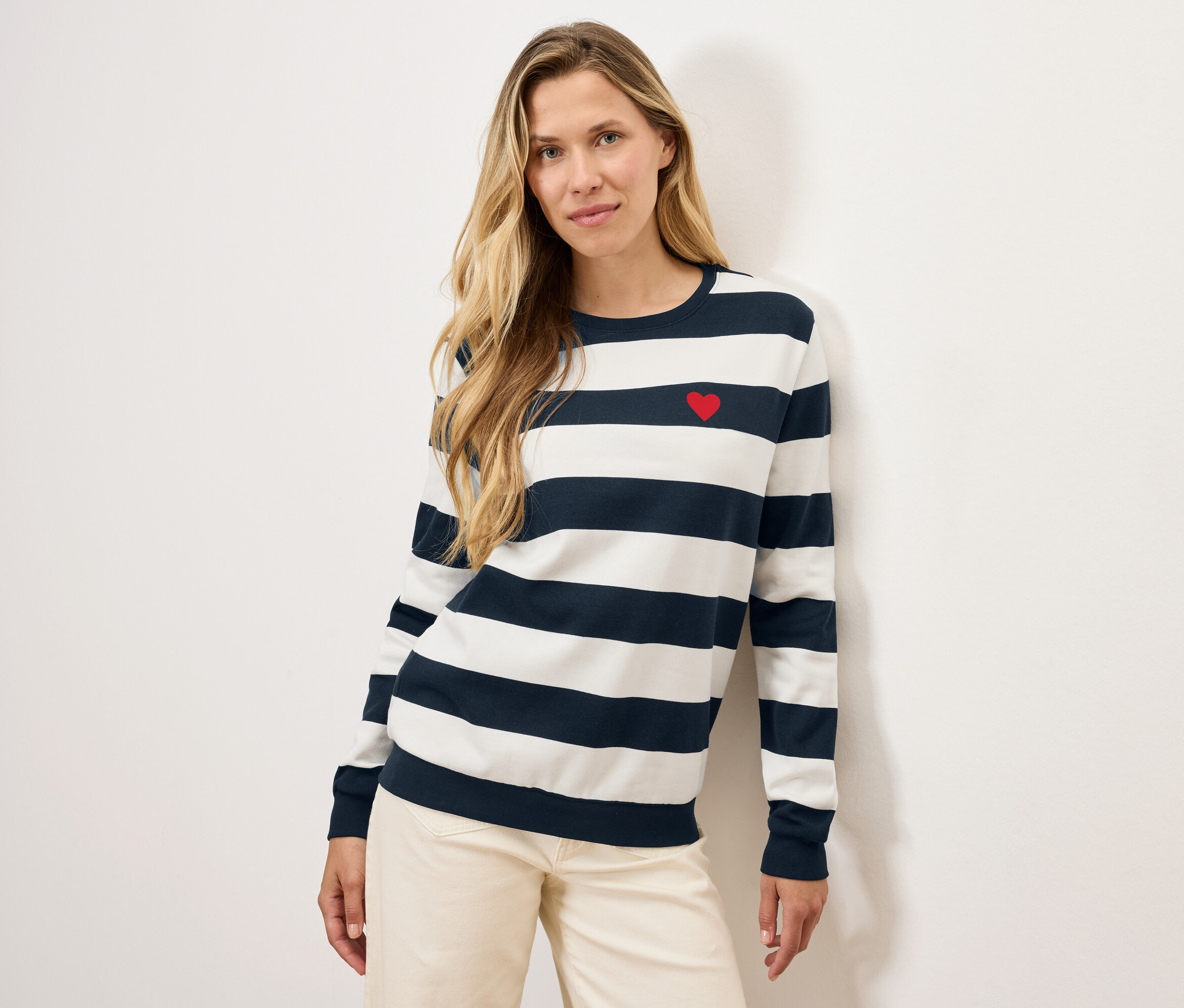 Blondýna pózuje v pruhovaném svetru s červeným srdcem a světlých kalhotách.