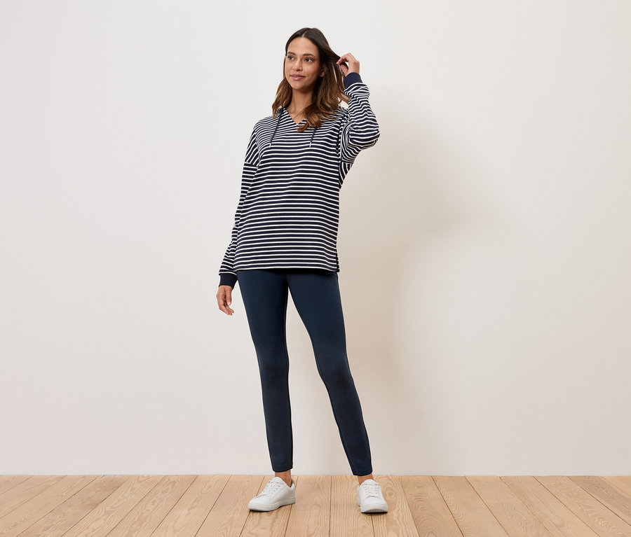 Žena stojí na dřevěné podlaze a má na sobě pruhovaný top, tmavě modré legíny a bílé tenisky.