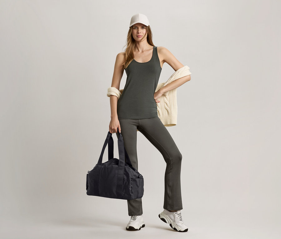 Žena v olivově zeleném topu a stahovacích sportovních kalhotách drží černou sportovní tašku.