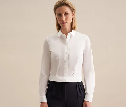 Žena v bílé halence slim fit Seidensticker s malým tmavým emblémem a tmavých skládaných kalhotách, stojí před světlým pozadím.