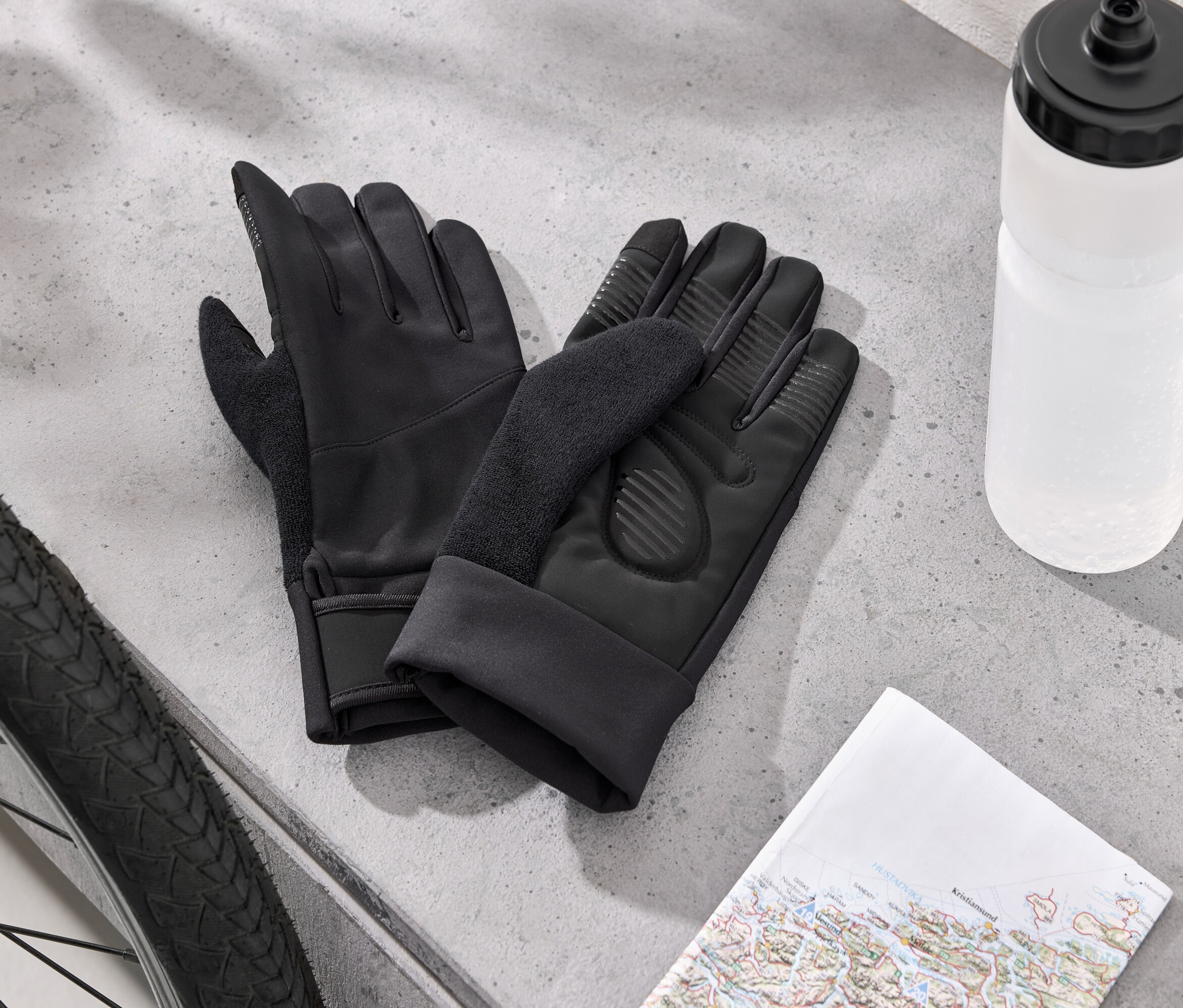 Dva černé cyklistické rukavice leží vedle pneumatiky kola, bílé láhve s vodou a mapy na šedém povrchu.