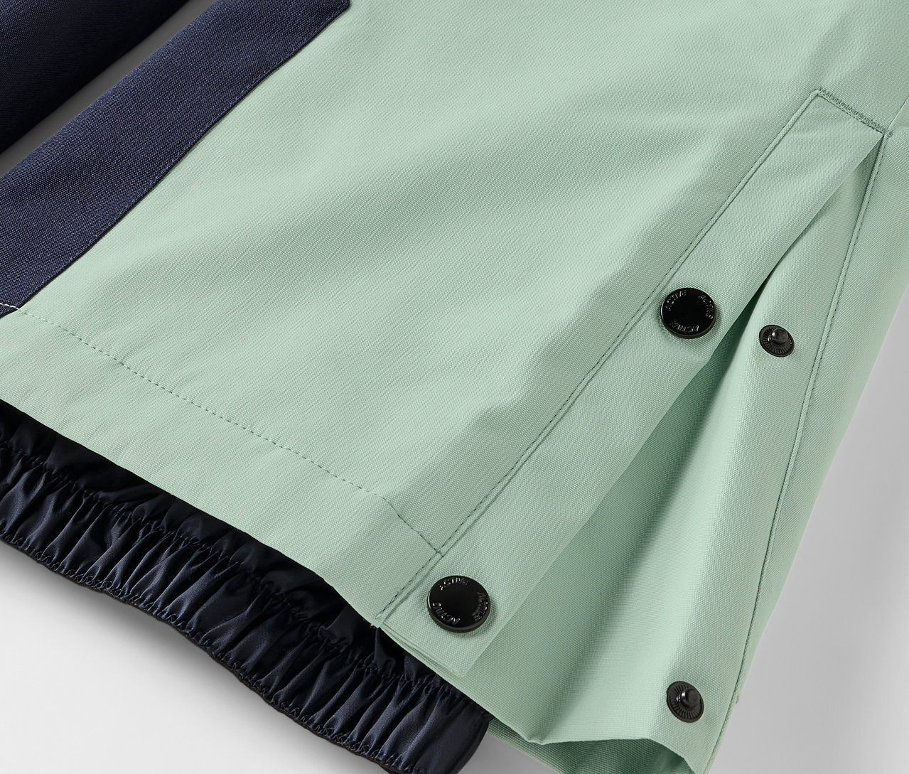 Detail světle zelených kalhot se zapínáním na knoflíky a elastickým lemem.