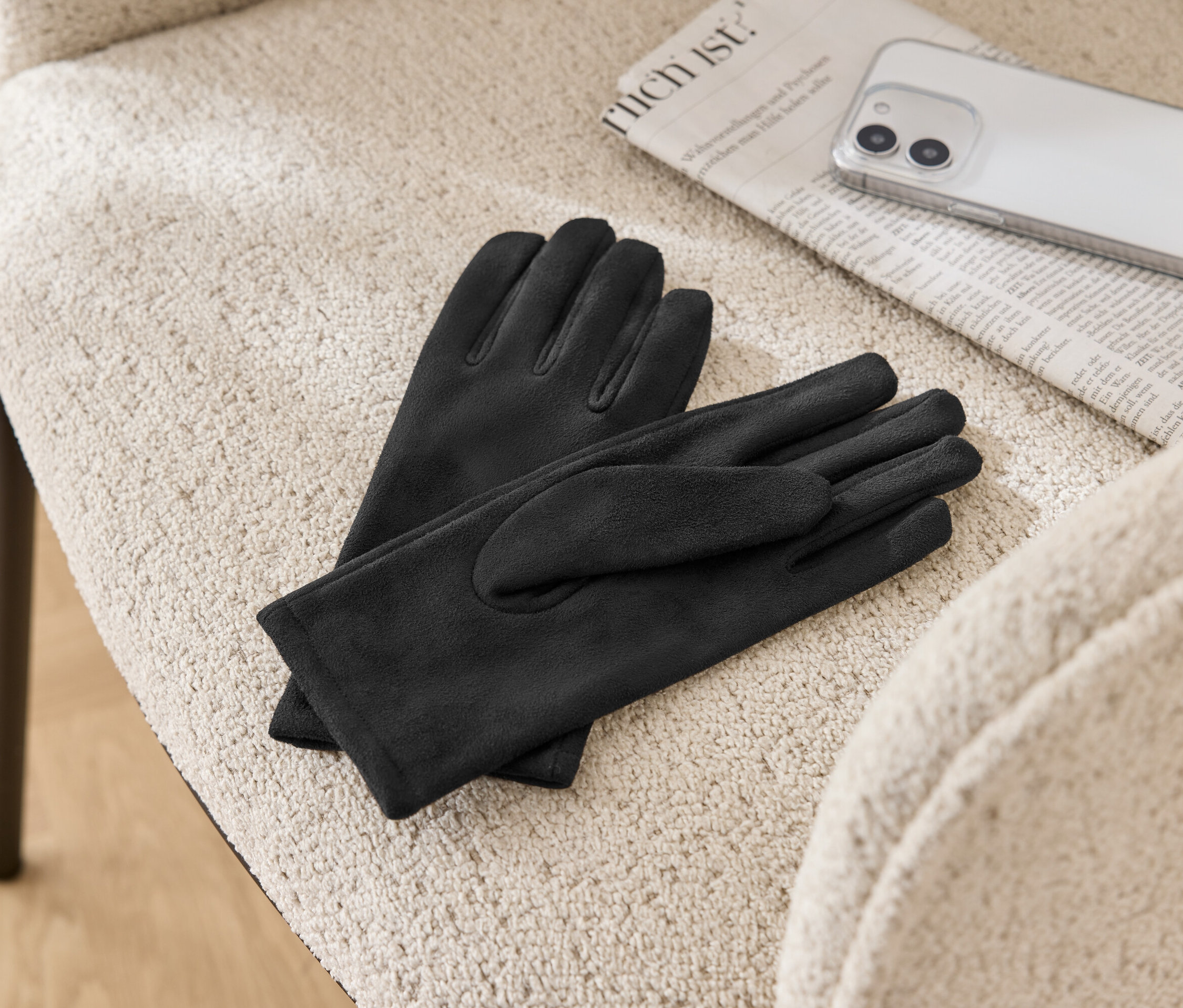 Na béžové židli leží pár černých kožených rukavic, noviny a mobilní telefon.