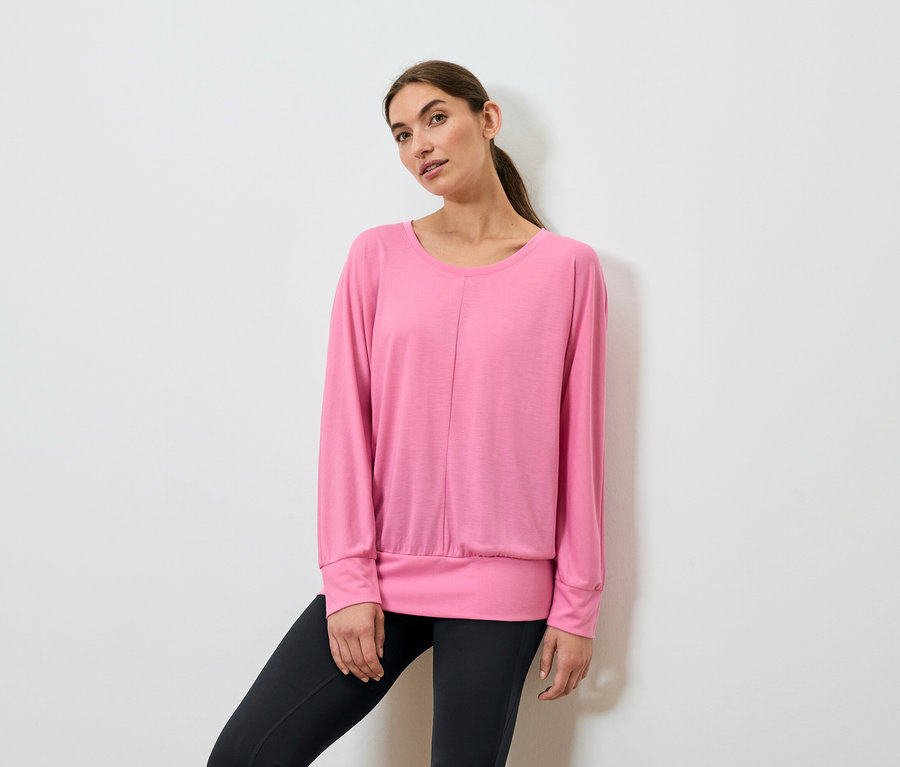Žena pózuje v růžovém sportovním topu s dlouhým rukávem a černých legínách.