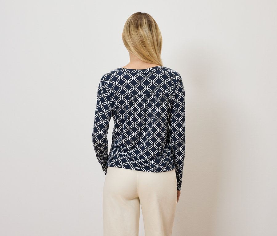 Žena zády k fotoaparátu má na sobě tmavě modré tričko s dlouhým rukávem a geometrickým vzorem a béžové kalhoty.