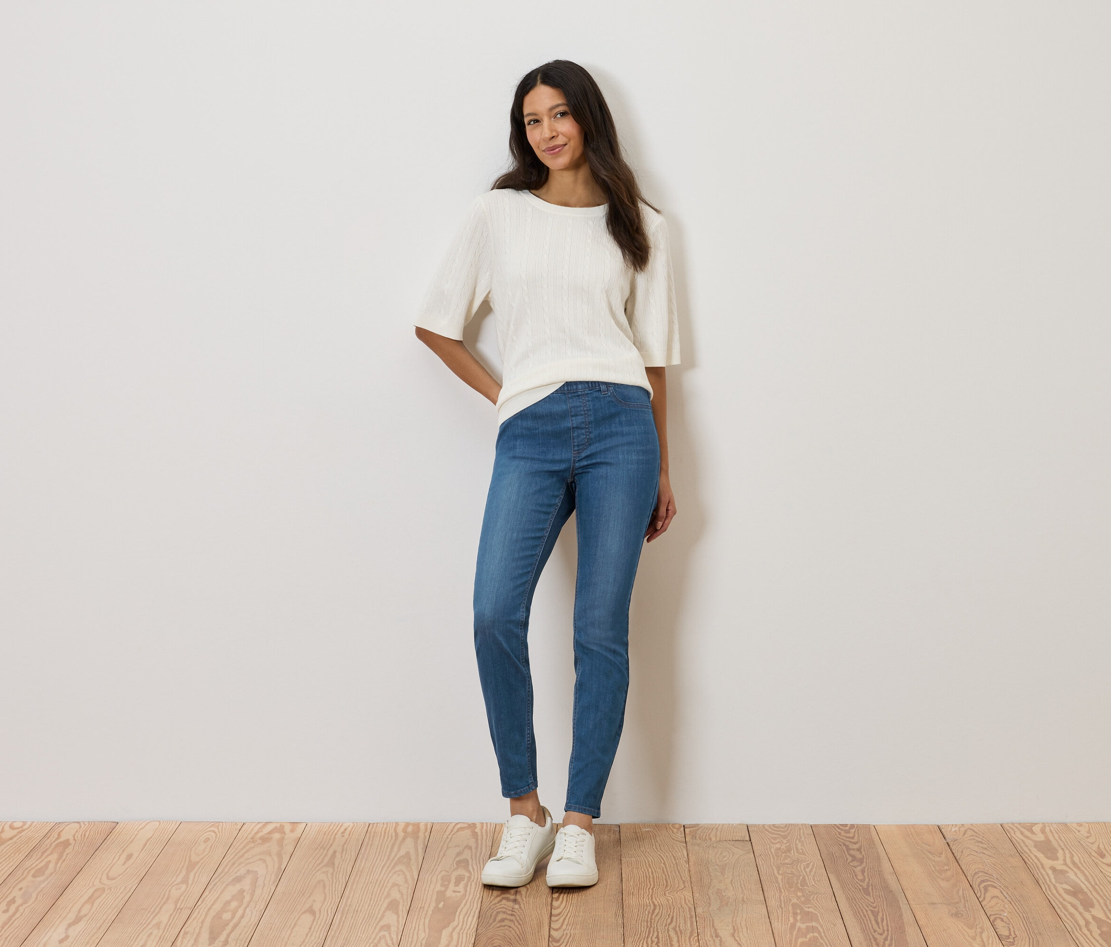 Žena stojí u zdi a má na sobě bílý top, modré džíny a bílé boty.