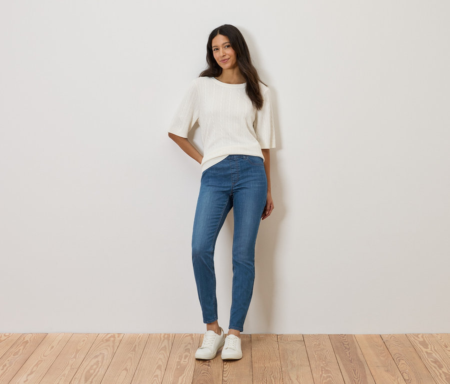 Žena stojí u zdi a má na sobě bílý top, modré džíny a bílé boty.